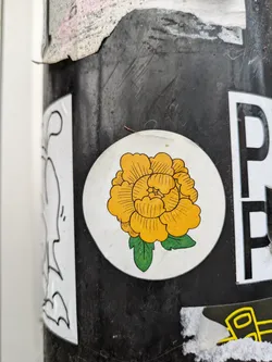Round white sticker featuring a stylized yellow flower with green leaves. The flower has a simple line drawing style.
