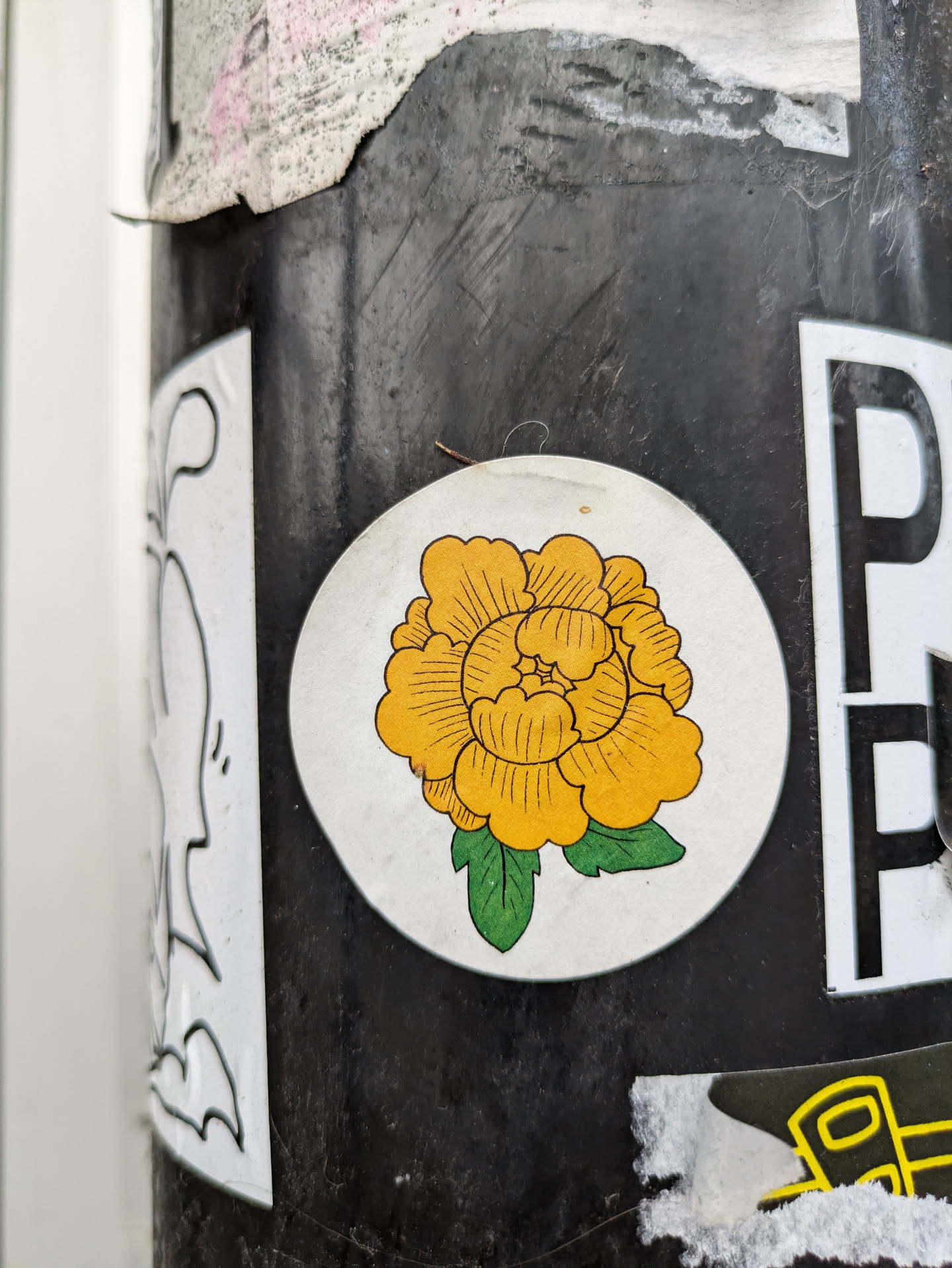 Round white sticker featuring a stylized yellow flower with green leaves. The flower has a simple line drawing style.