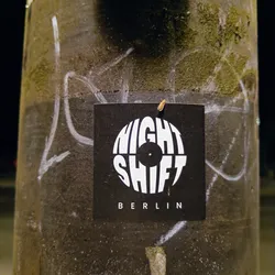 A black and white sticker with the text "NIGHT SHIFT" arranged in a circle resembling a vinyl record.  Below the circle, it says "BERLIN" in smaller font. It's affixed to a weathered, dark-colored pole.