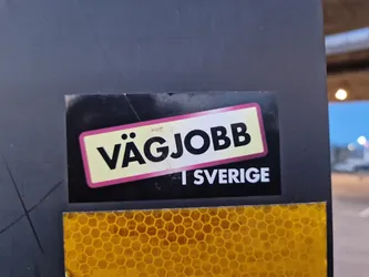 A black and yellow sticker with the text "Vägjobb i Sverige" which translates to "Road Job in Sweden". It's stuck on a dark gray surface and has a reflective yellow honeycomb pattern below it.