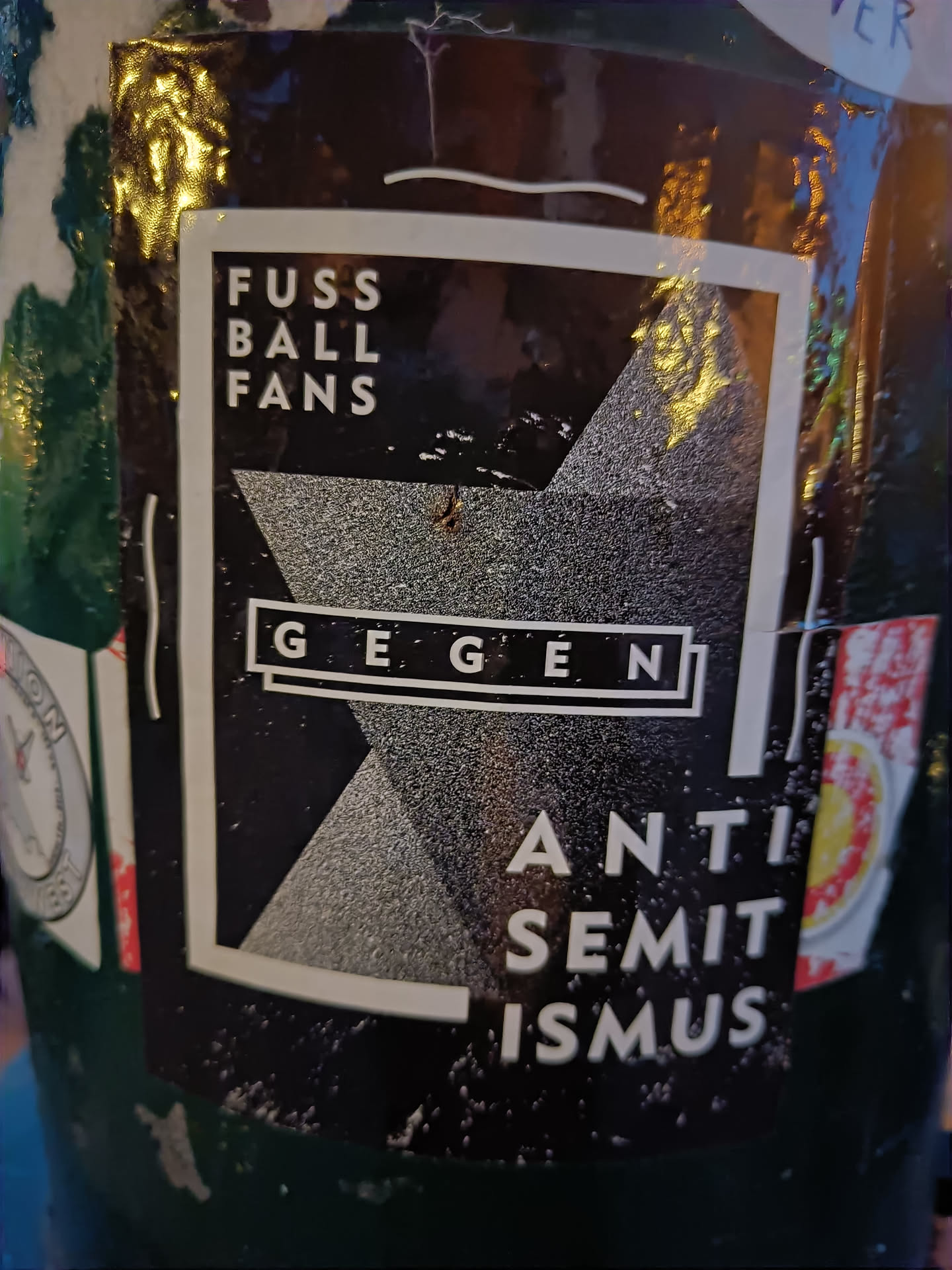 A black and white sticker with the text "Fussball Fans gegen Antisemitismus" (Football Fans against Antisemitism) in German. The design includes a geometric shape.