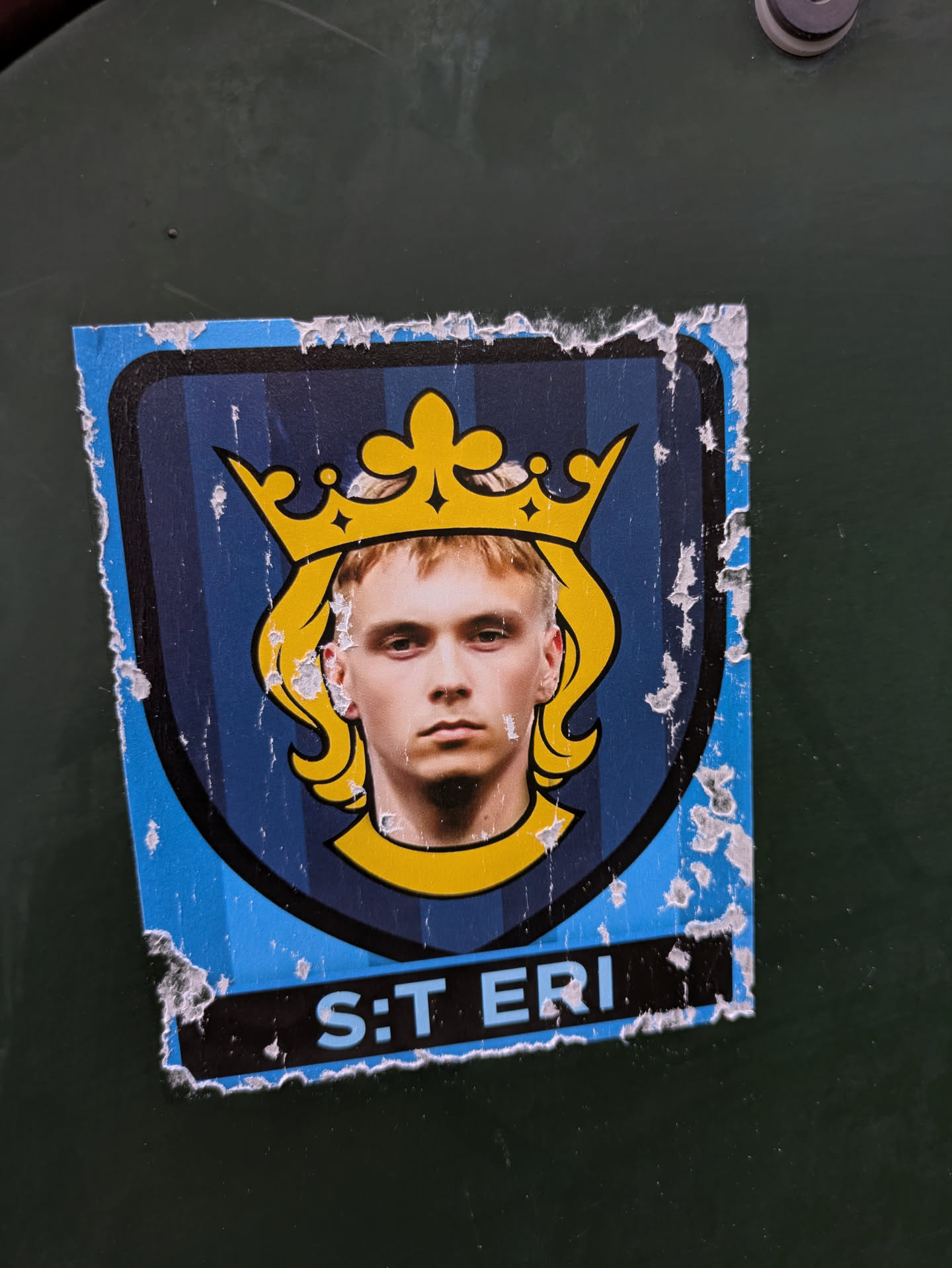 The sticker features a stylized shield with vertical dark blue stripes. At the top center is a prominent golden crown with intricate details. Within the shield, a young man with blond hair and a serious expression is depicted, his head framed by the crown and a yellow outline. Below the portrait, a black band contains the text 'S:T ERI' in light blue. The edges of the sticker are worn and torn, with visible peeling and white paper backing.