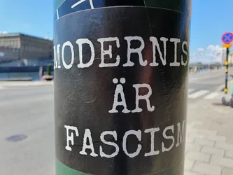 A black sticker with white text on a pole. The text says "Modernism är Fascism" in a typewriter-style font.
