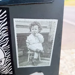 A grayscale sticker featuring a vintage photograph of a young child with curly hair sitting on a small stool. The child appears to be holding something small to their mouth. The photo has a slightly worn or aged look, adding to its nostalgic charm.