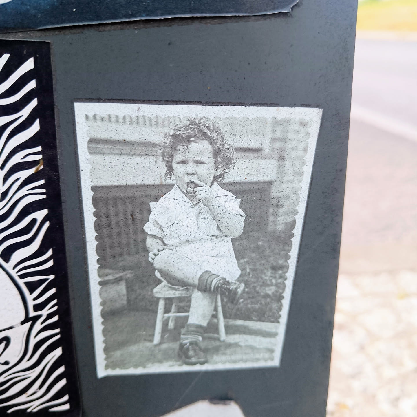 A grayscale sticker featuring a vintage photograph of a young child with curly hair sitting on a small stool. The child appears to be holding something small to their mouth. The photo has a slightly worn or aged look, adding to its nostalgic charm.