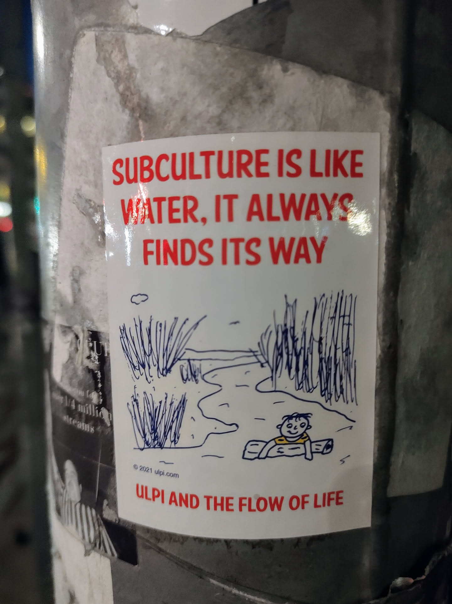 A white sticker with red text and a simple illustration of a person on a log in a river. The text on the sticker says, "Subculture is like water, it always finds its way."  At the bottom it says, "Ulpi and the flow of life". The illustration is a simple drawing of a waterway with reeds on either side, and a figure floating on a log.