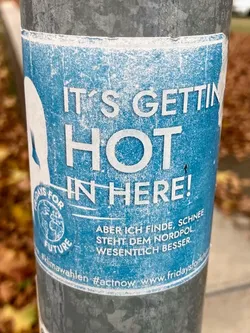 A blue sticker with the text "IT'S GETTING HOT IN HERE!" in large white letters. Below it, in smaller text, is a German phrase: "ABER ICH FINDE, SCHNEE STEHT DEM NORDPOL WESENTLICH BESSER."  The sticker also features a logo for Fridays For Future and social media hashtags and website address.