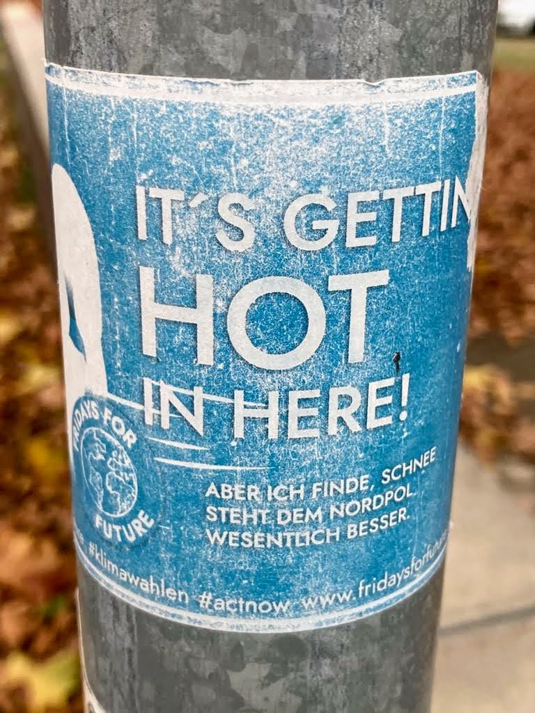 A blue sticker with the text "IT'S GETTING HOT IN HERE!" in large white letters. Below it, in smaller text, is a German phrase: "ABER ICH FINDE, SCHNEE STEHT DEM NORDPOL WESENTLICH BESSER."  The sticker also features a logo for Fridays For Future and social media hashtags and website address.