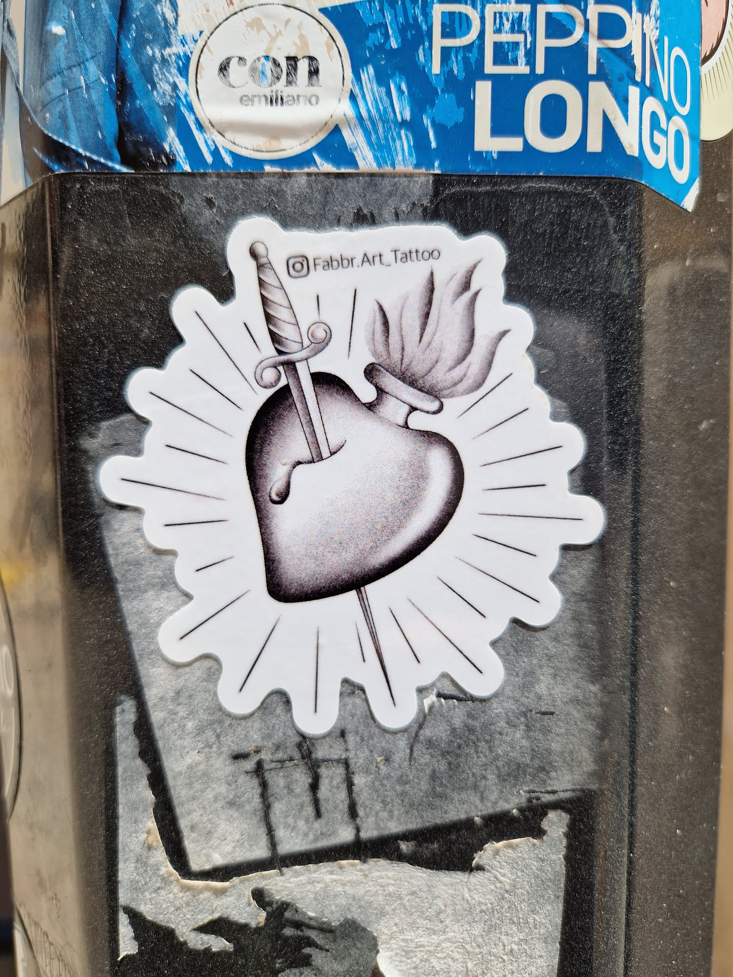 Black and white sticker with a design of a sacred heart pierced by a dagger with a flame on top. The sticker has a sunburst effect around it and the artist's Instagram handle is mentioned on it.