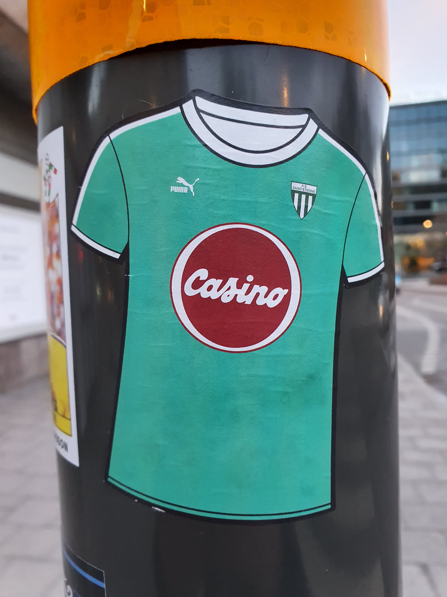 A sticker of a teal Puma jersey with a red circle on the chest that says Casino in a vintage-style font. The sticker is on a dark gray pole, in an urban setting.