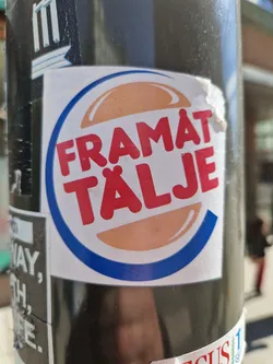 A sticker mimicking the Burger King logo but with the text "FRAMÄT TÄLJE".  The sticker is attached to a dark pole, partially obscuring other stickers underneath.