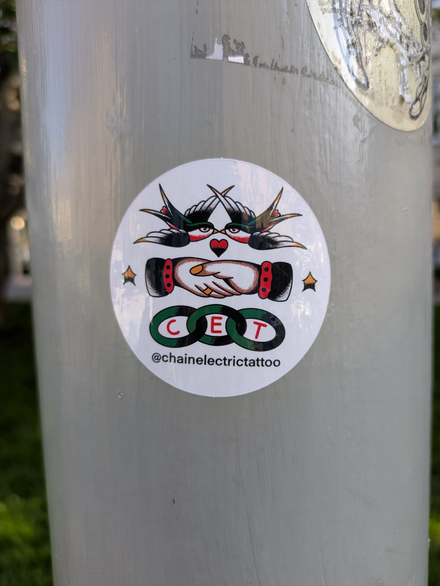 The sticker is circular with a white background. At the top, two stylized swallows in black and red with yellow accents are depicted in mid-flight, facing each other, with their wings extended. Between them, a red heart with black outlines is positioned. Below the swallows, two hands are shaking, rendered in a traditional tattoo style with detailed shading and a red cuff on one wrist. Encircling the center of the handshake are three interlocking rings, each with a green outline and a letter inside: 'C', 'E', and 'T'. Below the rings, the text '@chainelectrictattoo' is visible in black lettering. The overall aesthetic is reminiscent of traditional tattoo flash art.