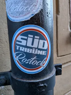 A circular sticker featuring a dark blue center with the words "SÜD TRIBÜNE" in bold, white, sans-serif capital letters at the top, and "Rostock" in a elegant, white script font at the bottom. The dark blue inner circle is framed by concentric rings of white, red, and a lighter blue, with a final thin white border. The sticker is adhered to a textured, black metal pole.