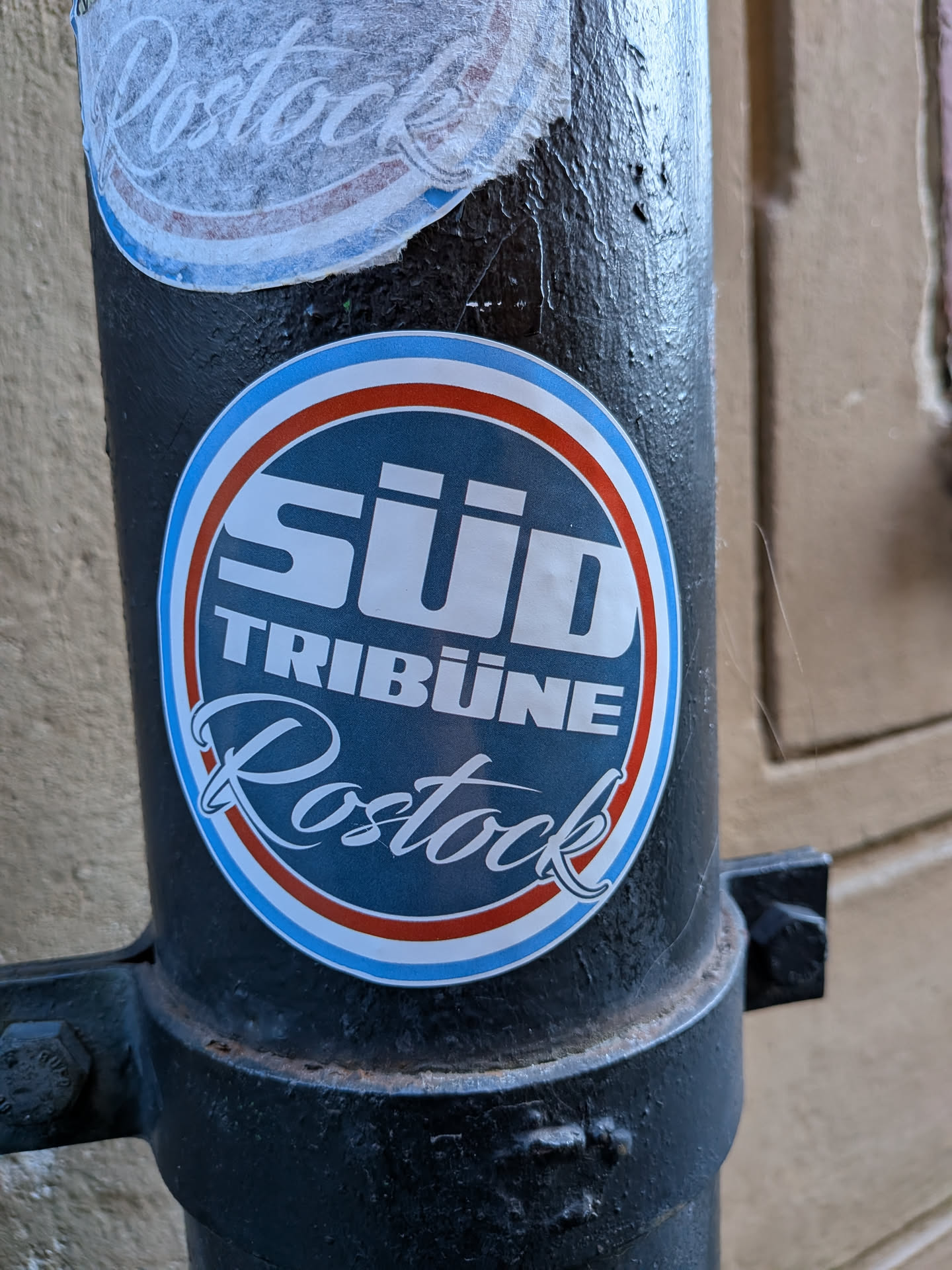 A circular sticker featuring a dark blue center with the words "SÜD TRIBÜNE" in bold, white, sans-serif capital letters at the top, and "Rostock" in a elegant, white script font at the bottom. The dark blue inner circle is framed by concentric rings of white, red, and a lighter blue, with a final thin white border. The sticker is adhered to a textured, black metal pole.