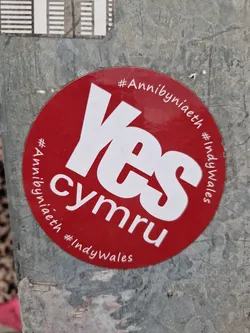 A red and white circular sticker advocating for Welsh independence. The sticker features the word 'Yes' prominently in white lettering against a red background.  Smaller text includes the hashtags #Annibyniaeth and #IndyWales, which are Welsh and English for 'Independence'.