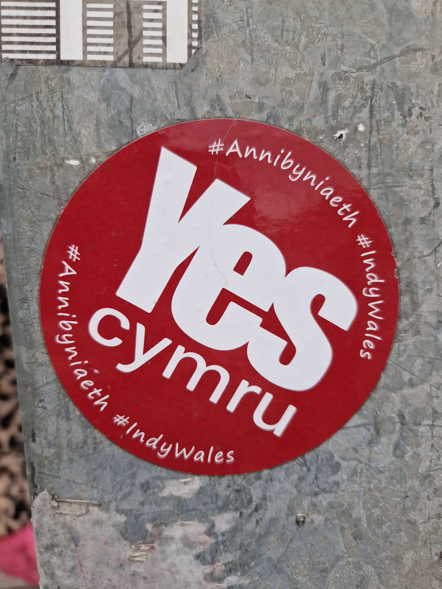 A red and white circular sticker advocating for Welsh independence. The sticker features the word 'Yes' prominently in white lettering against a red background.  Smaller text includes the hashtags #Annibyniaeth and #IndyWales, which are Welsh and English for 'Independence'.