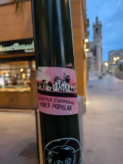 This sticker shows a line of black and white cartoon cats sitting together, with the words "HABITAR, COOPERAR, PODER POPULAR" printed in black on a pink background below them. It is affixed to a black pole.
