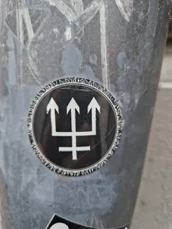 A round sticker with a black background and a white symbol resembling a trident or a stylized three-pointed star. The symbol is set against a decorative circular border.  The sticker is affixed to a gray surface which is marked with other graffiti or markings.