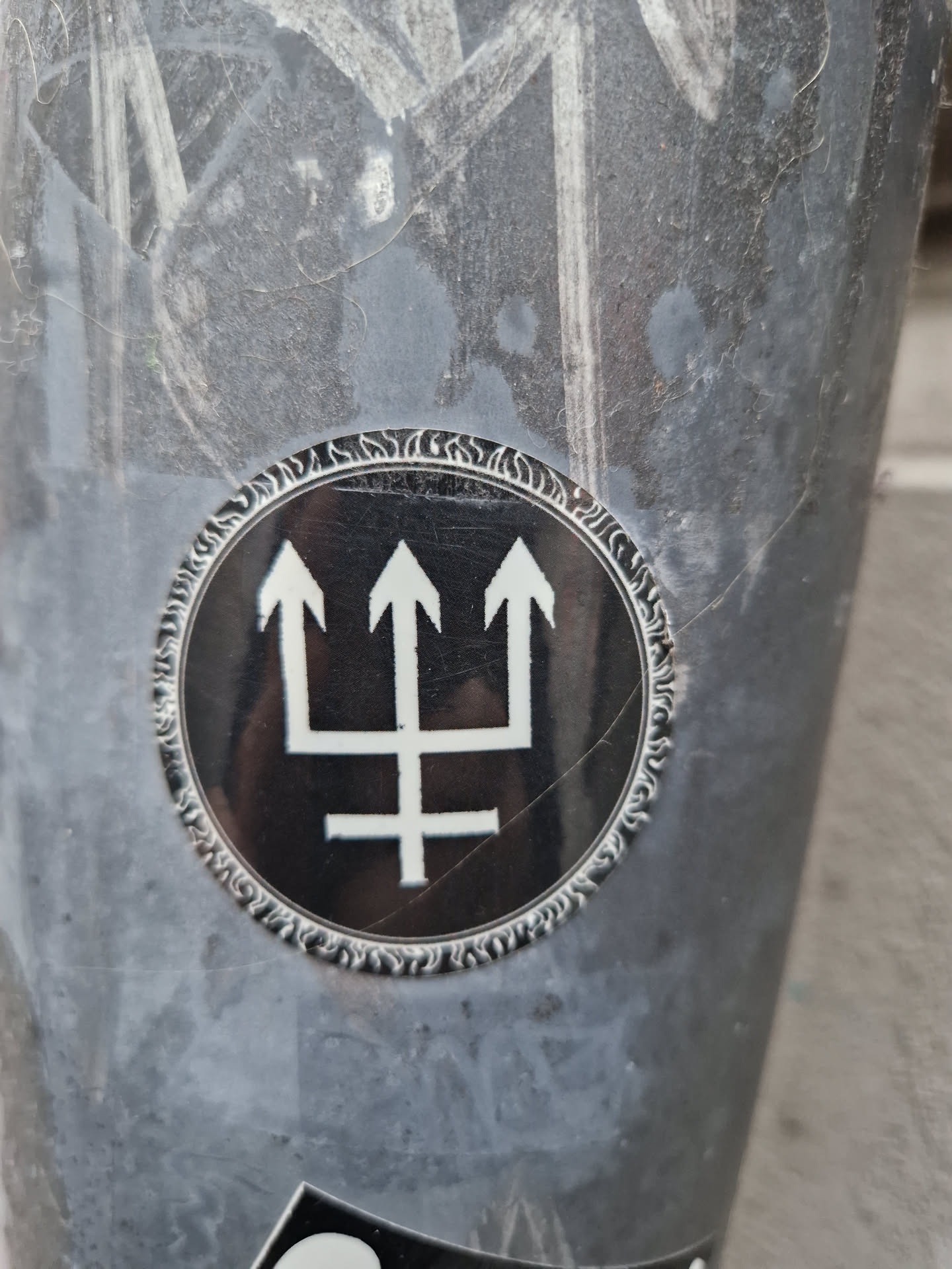 A round sticker with a black background and a white symbol resembling a trident or a stylized three-pointed star. The symbol is set against a decorative circular border.  The sticker is affixed to a gray surface which is marked with other graffiti or markings.