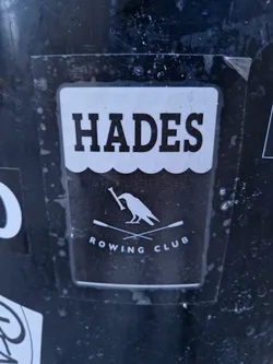 Black and white sticker featuring the name "HADES" in bold uppercase letters above an illustration of a crow holding a bone, crossed oars, and the words "ROWING CLUB" underneath. The design is simple and striking.