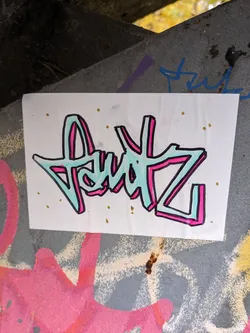 A sticker with a graffiti-style design. The main design is a stylized text "AuIZ" in light teal and pink with black outlines. It is set against a white background with some light golden dots scattered around. The sticker is placed on a grey surface with other graffiti visible around it.
