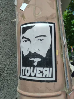 A black and white sticker featuring a stylized portrait of a man with a beard, the word 'TOVERAI' is printed underneath the portrait.  The sticker is affixed to a weathered pole.