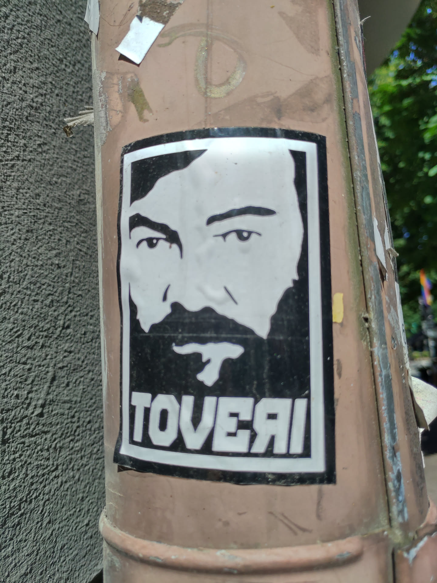 A black and white sticker featuring a stylized portrait of a man with a beard, the word 'TOVERAI' is printed underneath the portrait.  The sticker is affixed to a weathered pole.