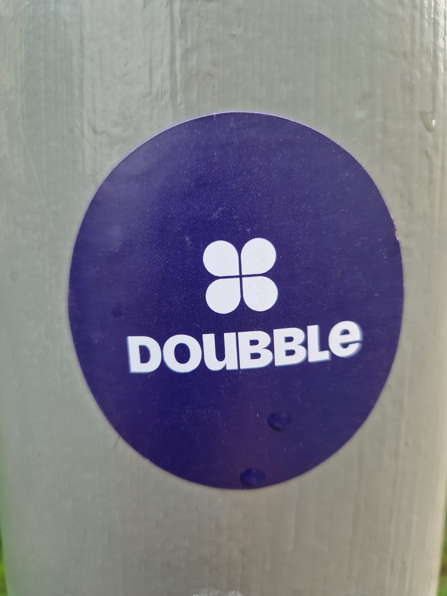 Round purple sticker with a white four-leaf clover and the word DOUBBLE in white uppercase letters.  The sticker is affixed to a light gray pole.