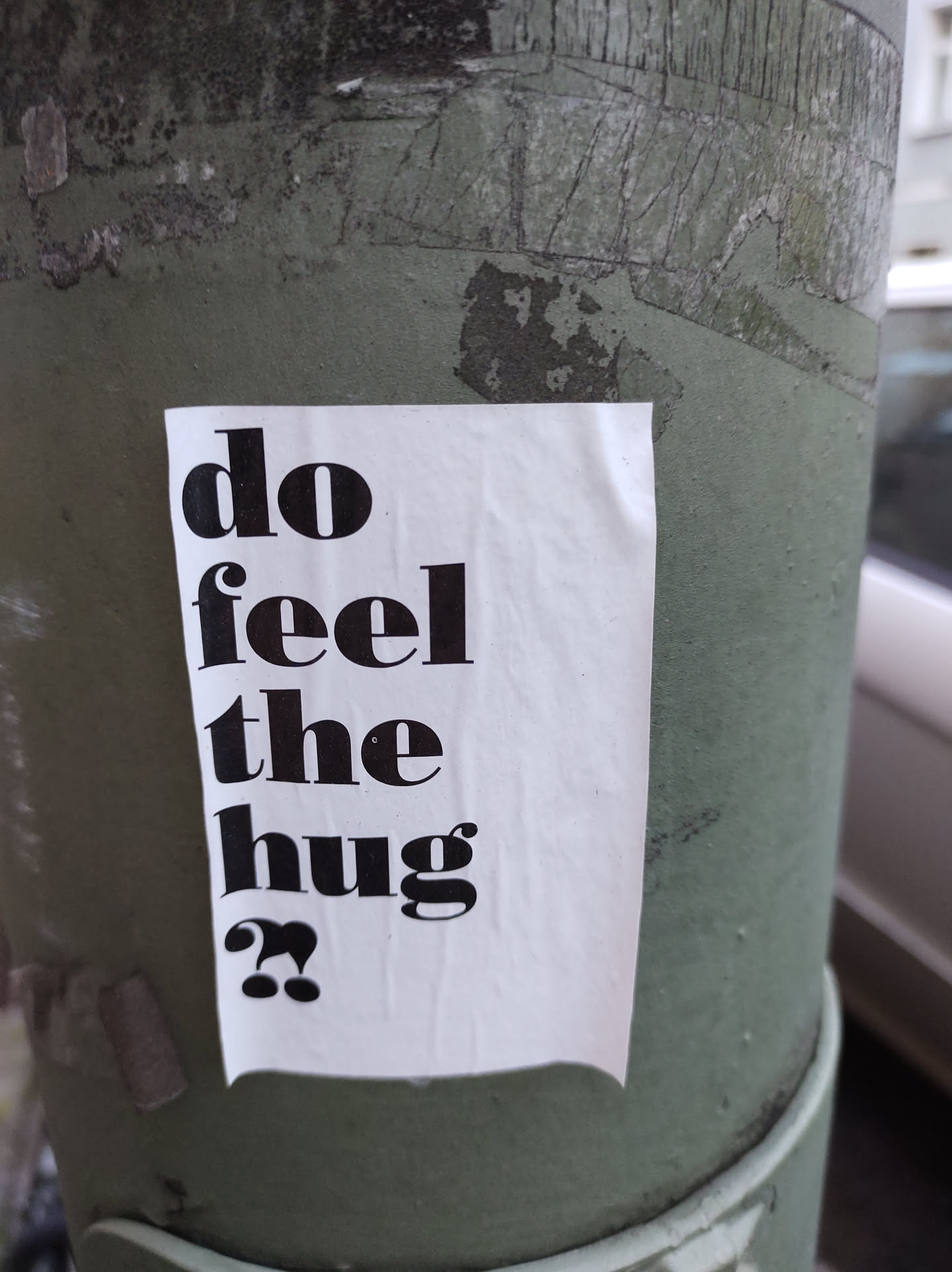 A white sticker with black text on a green pole. The text says: "do feel the hug??"