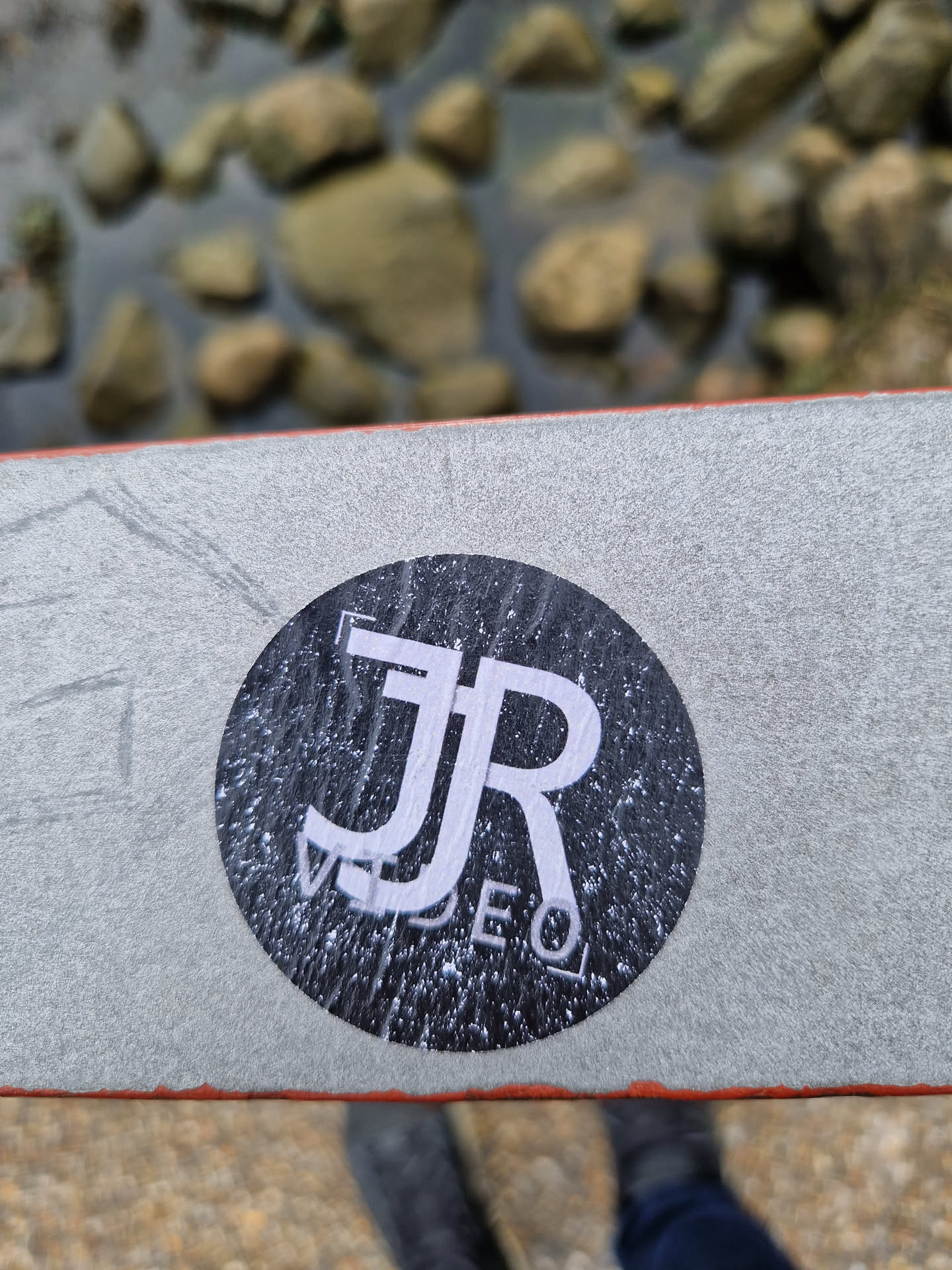A black and white round sticker with the logo 'JR VIDEO' is stuck on a grey surface. The sticker appears slightly worn or weathered, adding a cool, vintage touch.  The background is blurred, showing rocks near a body of water. 