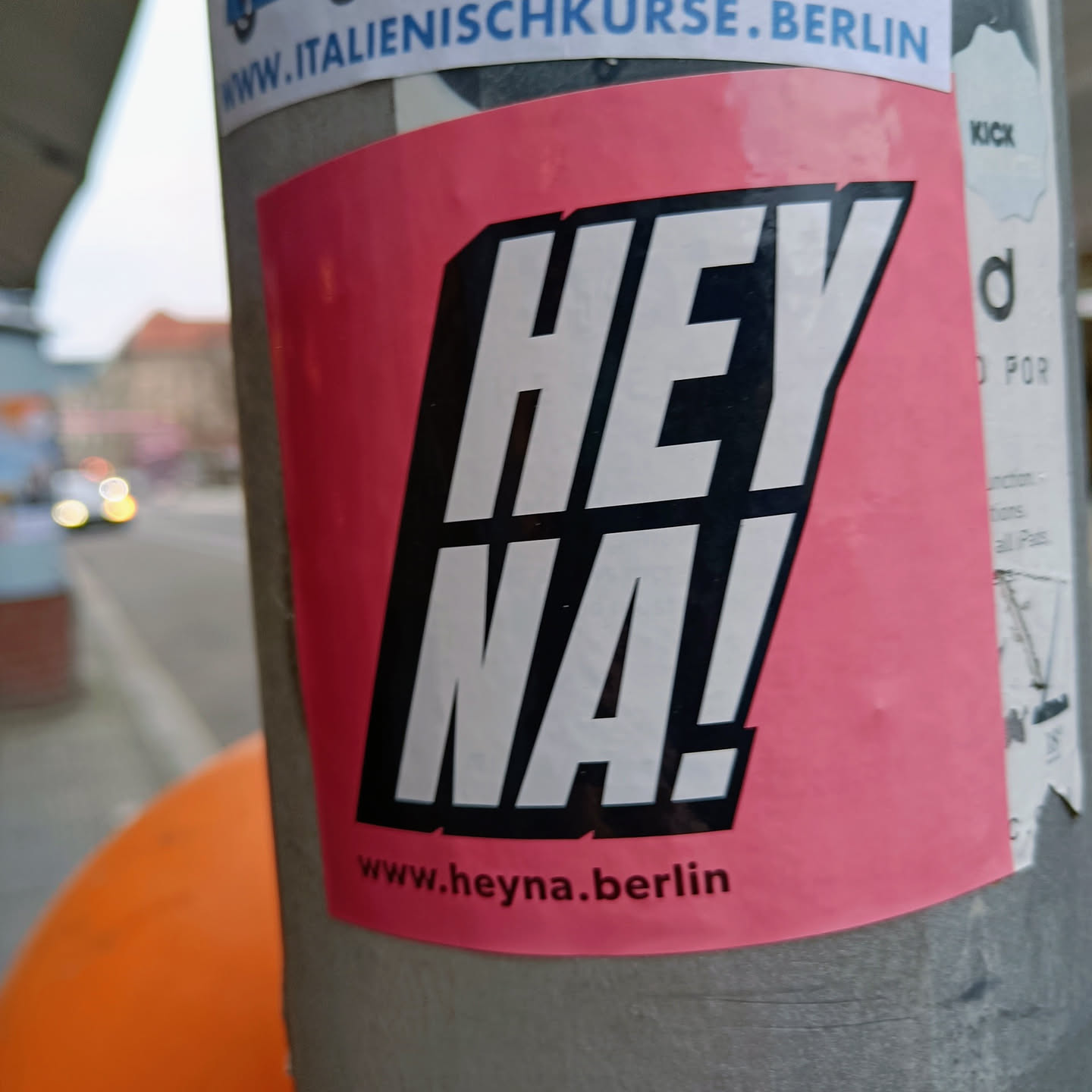 Pink and black sticker with the text 'Hey Na!' and the website www.heyNa.berlin. The sticker is placed on a grey pole.