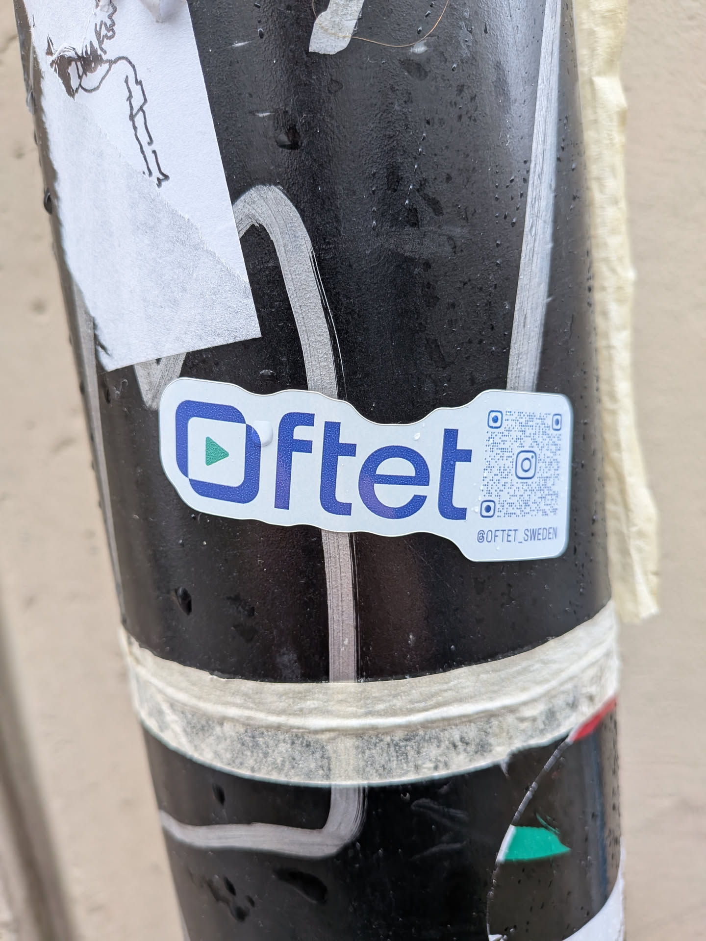 A white, irregularly shaped sticker with rounded corners, featuring the word 'Oftet' in dark blue sans-serif font. To the left of 'Oftet' is a dark blue square outline containing a vibrant green play button triangle. To the right of the text is a QR code, and below it, the Instagram handle '@OFTET_SWEDEN' is printed in smaller dark blue text. The sticker is applied to a dark, possibly wet, textured pole, with some white tape visible above and below it.