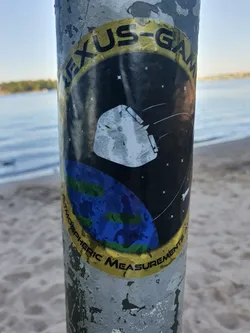 A sticker featuring a design resembling a space mission patch.  It shows a spacecraft orbiting a stylized Earth against a dark background. The text 'NEXUS-GAM Atmospheric Measurements' is prominently displayed in a circular band around the main image.