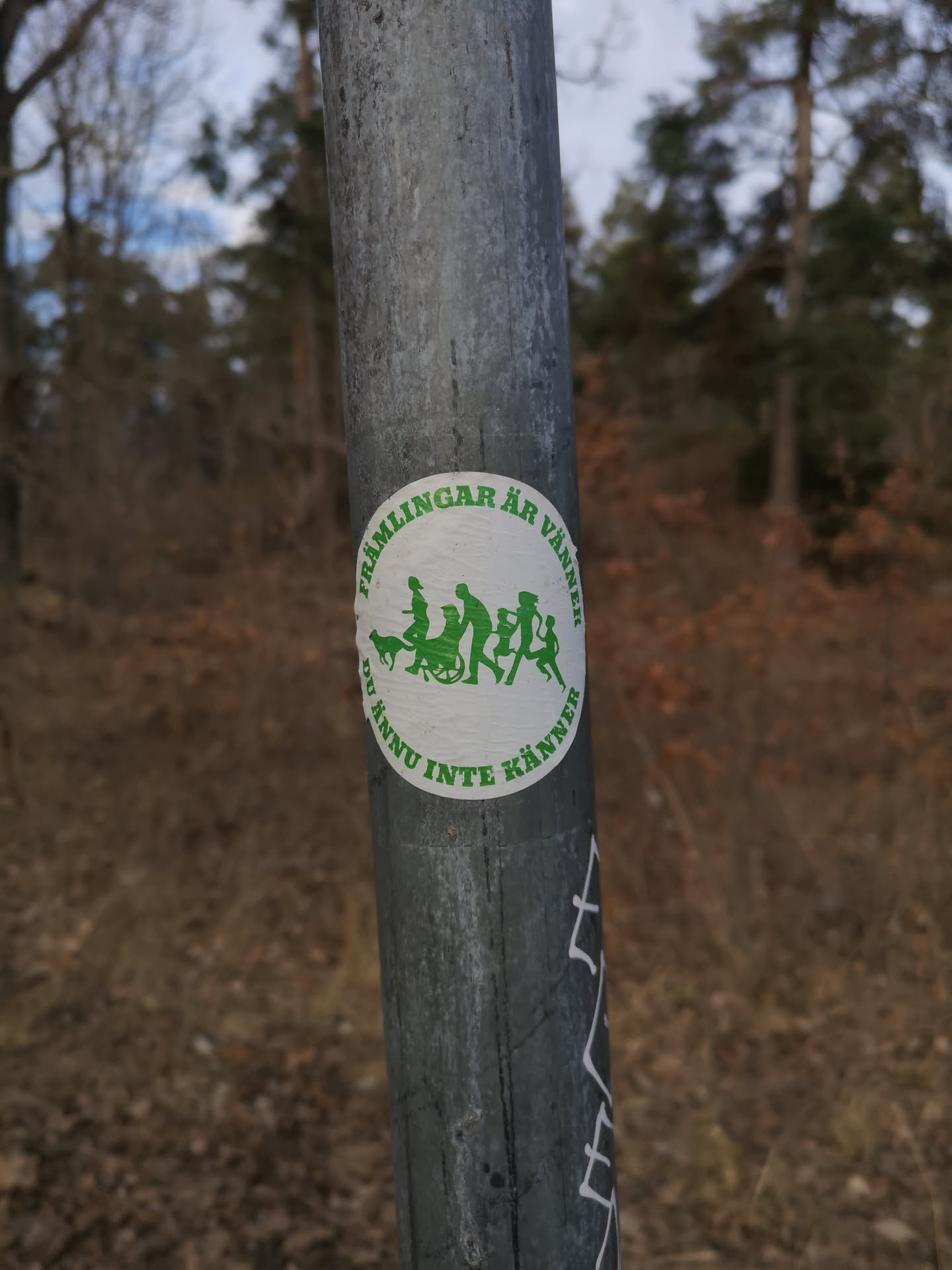 A circular white sticker featuring green silhouettes of diverse figures walking together. The group includes a dog, several adults, one of whom is in a wheelchair, and a child. The figures are stylized and appear to be in motion. Encircling the central image is green text in Swedish. The sticker is affixed to a weathered, grey metal pole, showing some minor surface imperfections and wrinkles.