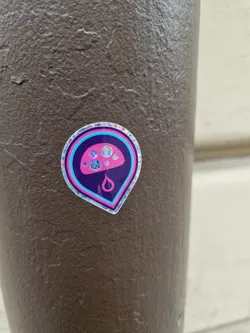 A holographic sticker featuring a pink mushroom with a droplet underneath. It has a map pin shape and is stuck on a brown surface.