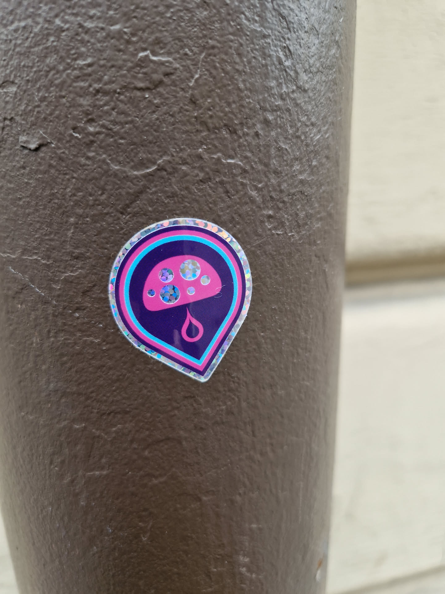 A holographic sticker featuring a pink mushroom with a droplet underneath. It has a map pin shape and is stuck on a brown surface.