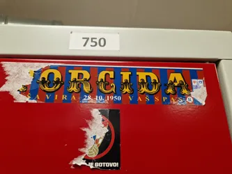 A worn-out sticker featuring the word "ORCIDA" in a vintage circus font. Below the main text, it says "SAVIRA 28.10.1950 VAŠ PAS", which might refer to a date and a pass. The sticker has a red, blue, and yellow color scheme. Part of a lower sticker is visible, with some text readable as "JE GOTOVO!"