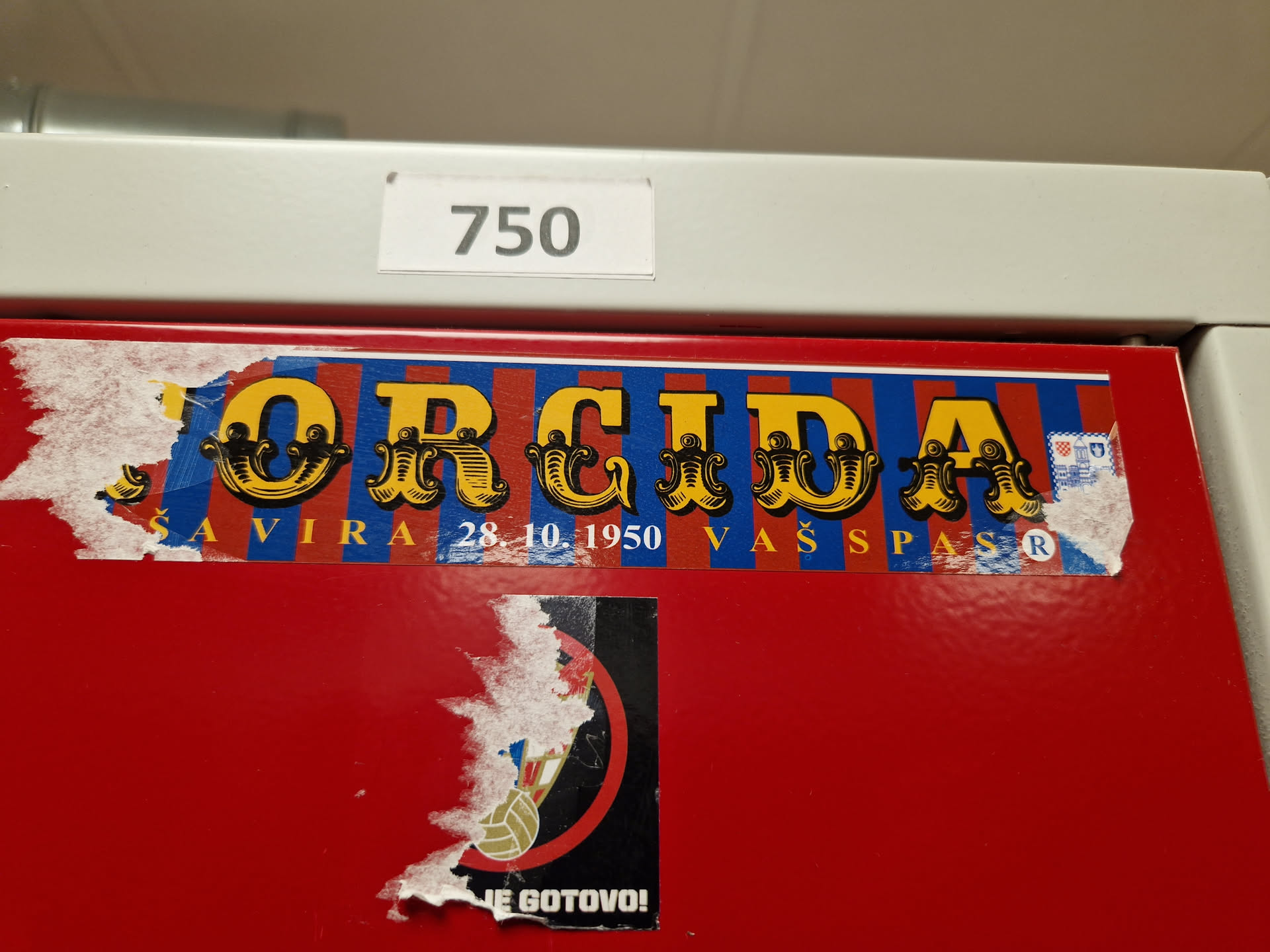 A worn-out sticker featuring the word "ORCIDA" in a vintage circus font. Below the main text, it says "SAVIRA 28.10.1950 VAŠ PAS", which might refer to a date and a pass. The sticker has a red, blue, and yellow color scheme. Part of a lower sticker is visible, with some text readable as "JE GOTOVO!"