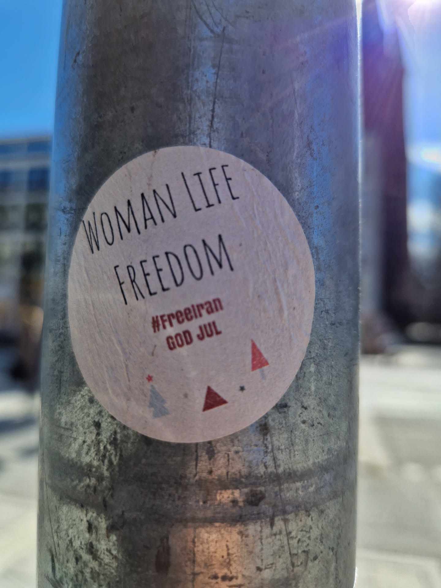 A round sticker on a metal pole with the text "Woman Life Freedom" at the top and the hashtags "#FreeIran" and "God Jul" (Merry Christmas in Swedish) at the bottom. There are also small illustrations of Christmas trees.
