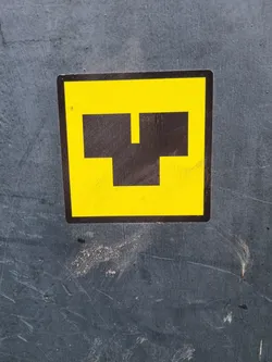 A yellow square sticker with a black abstract symbol resembling a stylized letter 'T' or a simple machine part. The sticker is affixed to a dark gray surface showing signs of wear and tear.