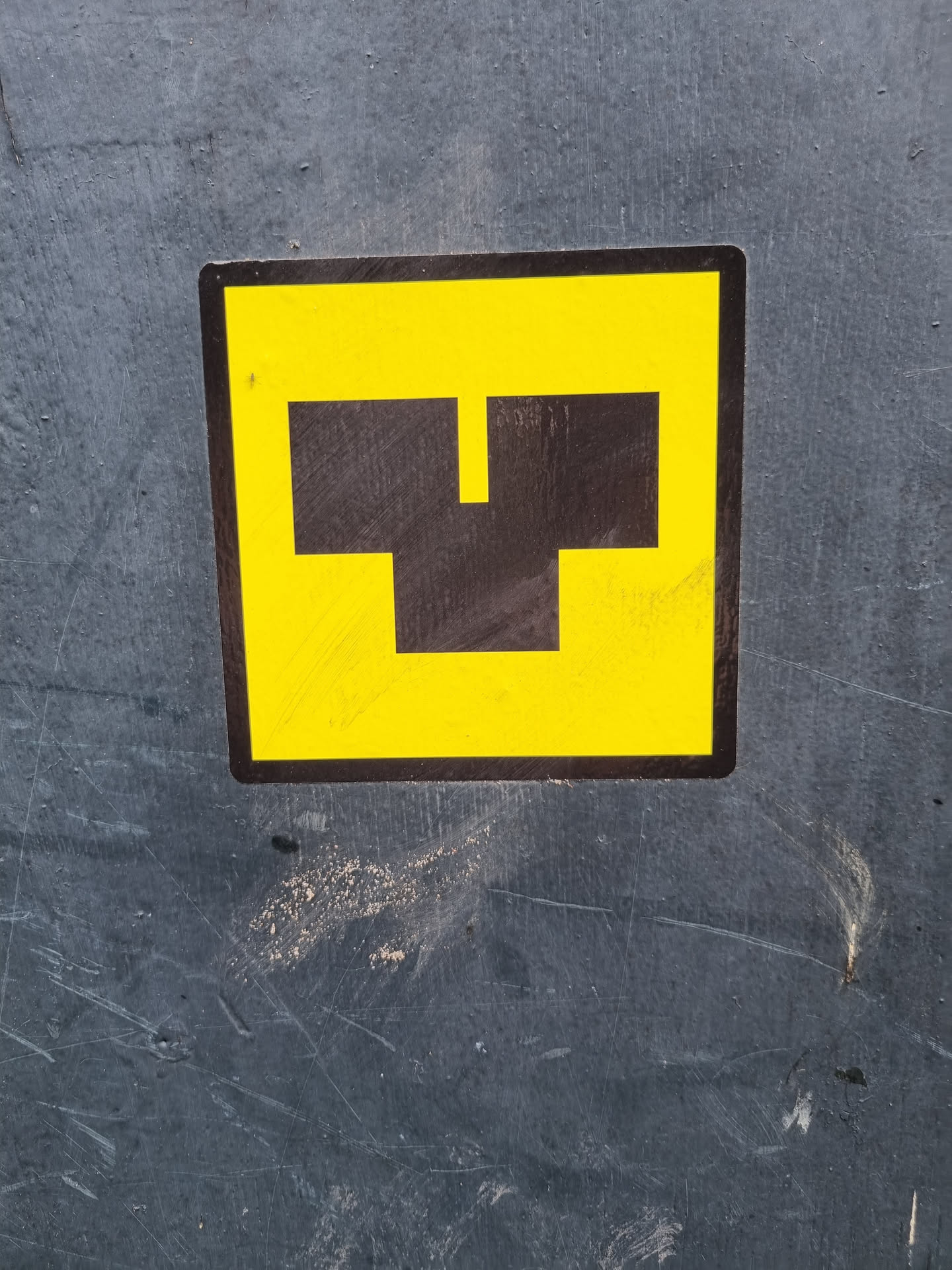A yellow square sticker with a black abstract symbol resembling a stylized letter 'T' or a simple machine part. The sticker is affixed to a dark gray surface showing signs of wear and tear.