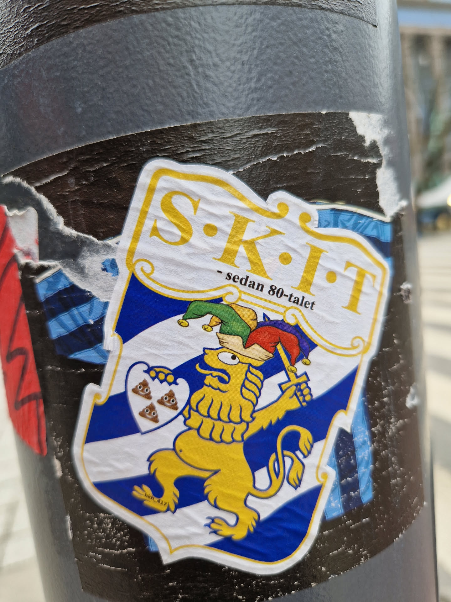 A sticker featuring a yellow lion holding a sword, standing on a blue and white shield with three poop emojis. The text 'SKIT - sedan 80-talet' is written at the top. The style is humorous and slightly irreverent.