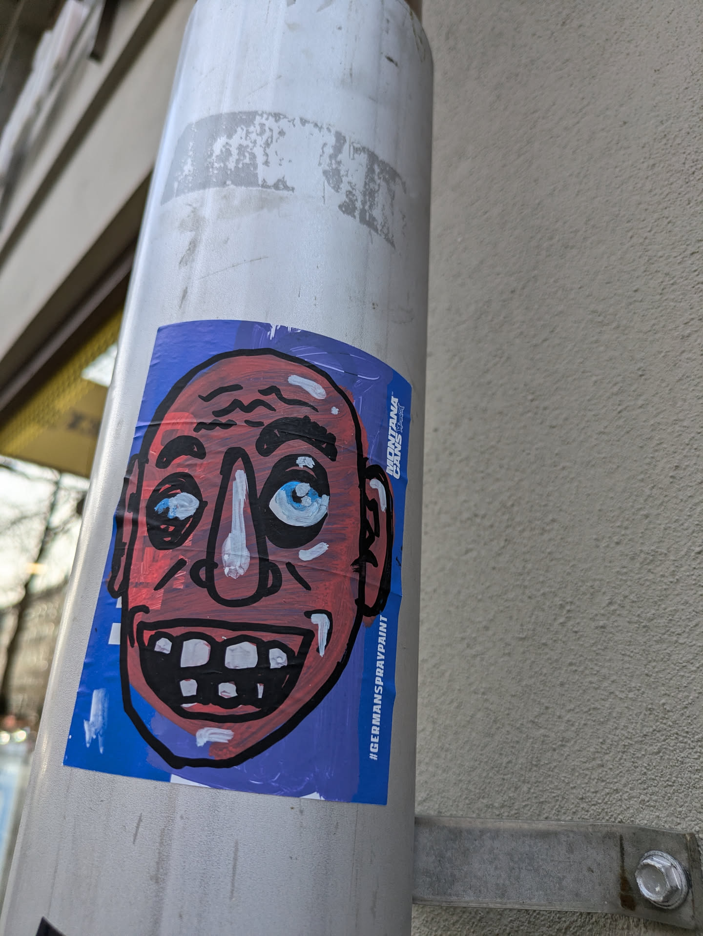 A vibrant sticker featuring a cartoonish bald man with a wide, toothy grin. The style is reminiscent of graffiti art, with bold outlines and slightly uneven coloring. The background is a solid blue, providing a contrast to the character's reddish skin tone.  The sticker also contains the text '#GERMANSPRAYPAINT'.