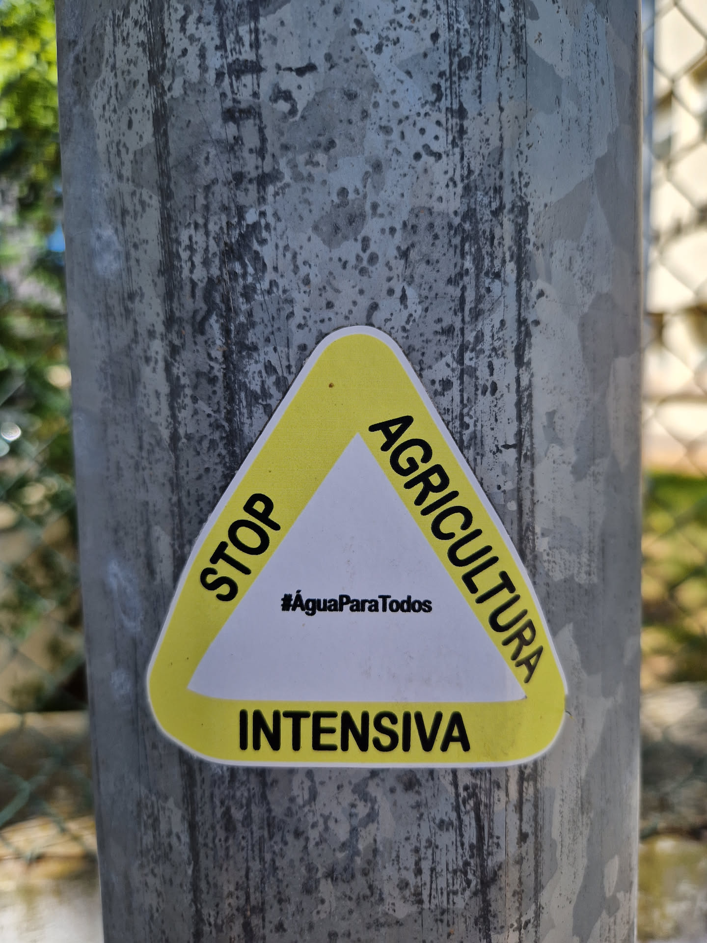 Triangular yellow sticker with the text "STOP AGRICULTURA INTENSIVA" and the hashtag #AguaParaTodos in the center. The sticker is attached to a gray metal pole.