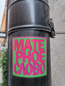 A sticker with the text "MATE PHOE CΛOBO" in pink letters on a green background. The sticker is affixed to a dark-colored pole.
