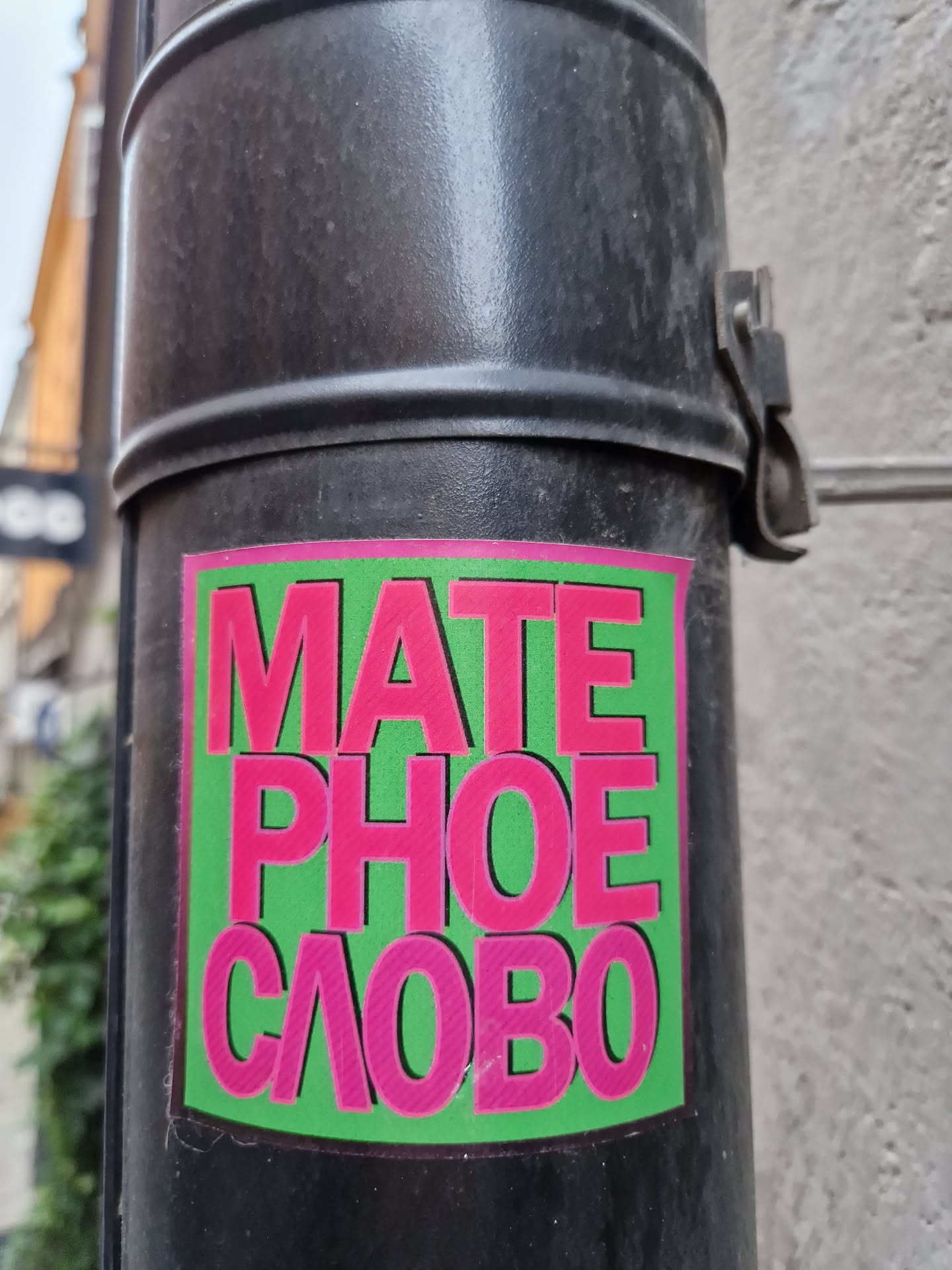 A sticker with the text "MATE PHOE CΛOBO" in pink letters on a green background. The sticker is affixed to a dark-colored pole.