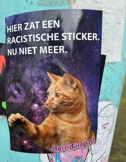 A sticker featuring an orange cat in space, with text in Dutch saying "Here was a racist sticker. Now not anymore."  The image also includes a hammer and sickle symbol.  The website derodelap.nl is visible in the bottom-right corner.