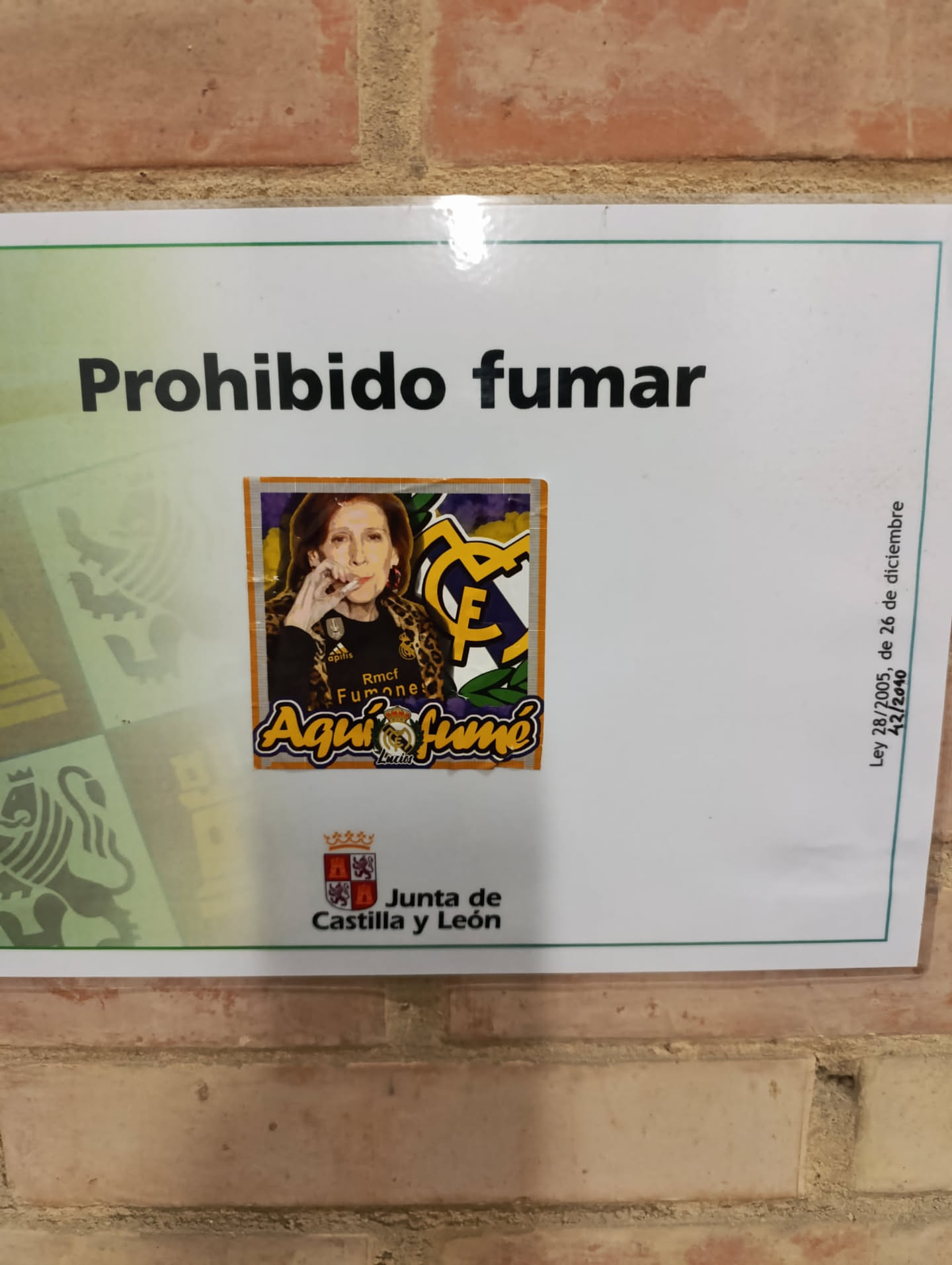 The sticker is a rectangular, digitally printed image with a slightly uneven orange border, applied to a "Prohibido fumar" (No Smoking) sign. It features a stylized illustration of an older woman with short, reddish-brown hair, wearing a leopard print jacket over a dark top. She is depicted holding a lit cigarette to her lips, exhaling smoke, with a calm, almost defiant expression. On her dark top, small white text reads "apitis" and "Rmcf Fumone". To the right of the woman, a prominent yellow and purple "RMCF" logo, characteristic of Real Madrid C.F., is displayed. The background of the sticker is a vibrant mix of purple and yellow, with abstract patterns. Below the woman, the phrase "Aquí fumo" (Here I smoke) is written in large, yellow, bubble-style letters with an orange outline. Beneath "fumo", a small crown and a stylized lion figure are visible, with the tiny word "Lucios" below them. The overall style is illustrative and bold, creating a humorous and rebellious contrast with the underlying sign.