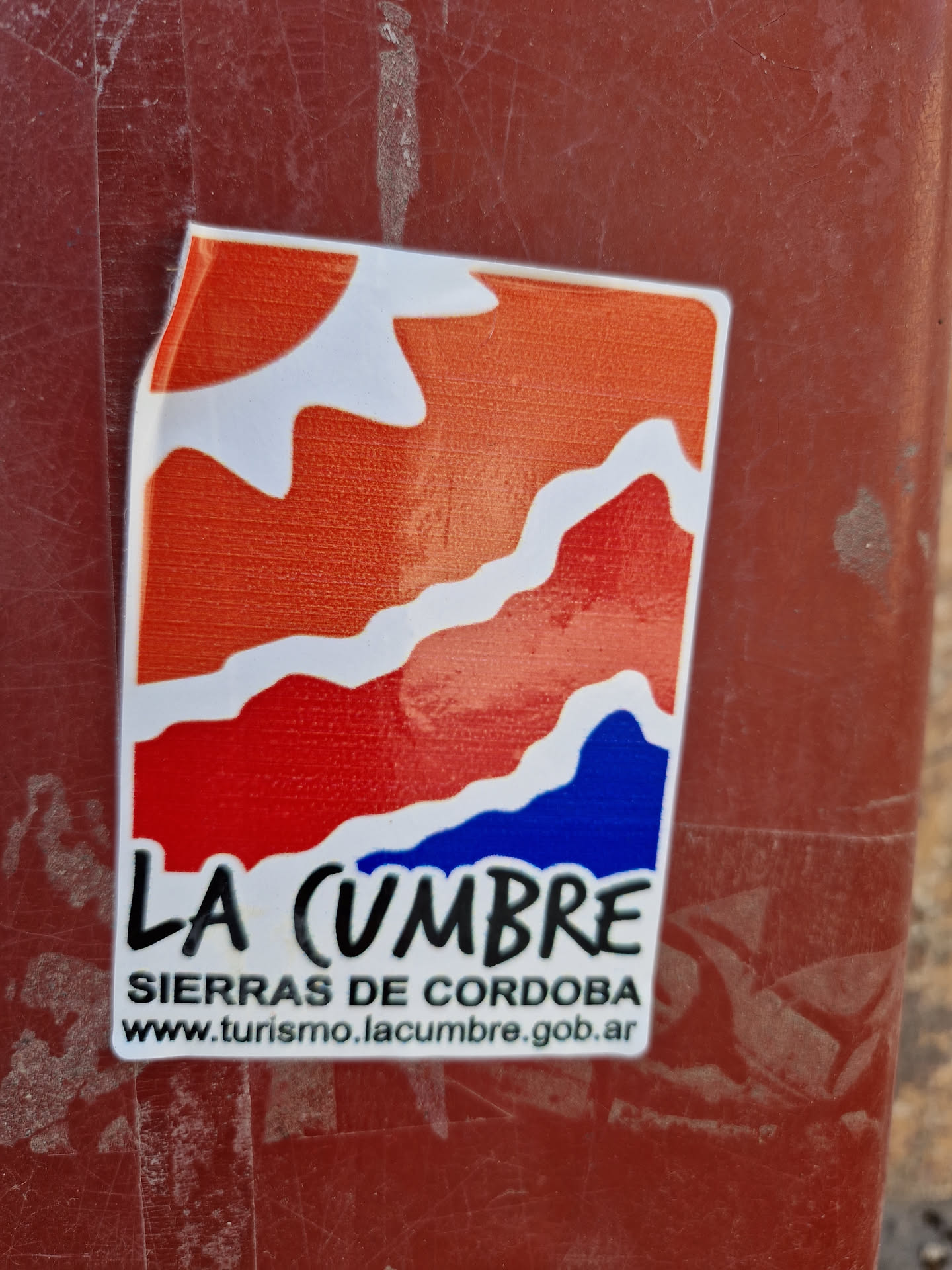A sticker featuring a stylized landscape design with a sun and mountains in orange, red and blue colors. Text on the sticker reads "LA CUMBRE" and "SIERRAS DE CORDOBA" with a website address below it. 