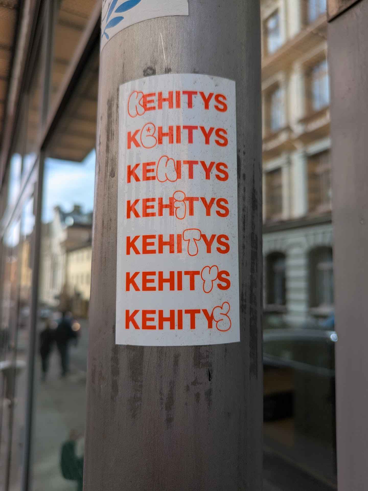 The sticker features the word "KEHITYS" printed in orange multiple times on a white background, affixed to a metal pole.